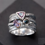 Large signet ring with crisscrossing textured black silver twigs and 3 stones on top. A pink triangle spinel, a round blue tourmaline, both set in white gold, and an oval purple tourmaline claw set in a red gold shawl
