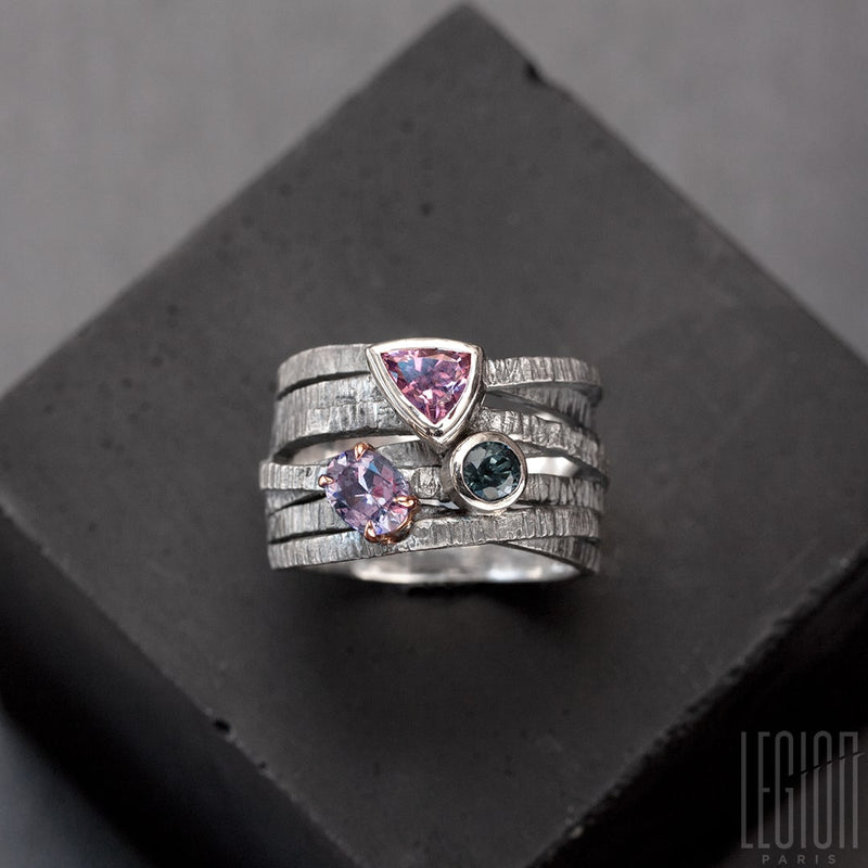 Large signet ring with crisscrossing textured black silver twigs and 3 stones on top. A pink triangle spinel, a round blue tourmaline, both set in white gold, and an oval purple tourmaline claw set in a red gold shawl