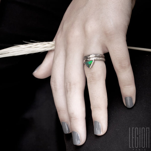 woman's hand wearing two white gold rings. A faceted wedding band and a ring composed of a textured band and a black opal with green and orange lights set in close on top. 