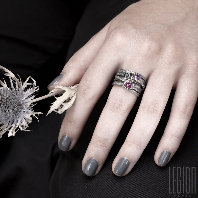 Woman's hand wearing a large Chevalière with textured and interlocking black silver twigs and 3 stones on top. A pink triangle spinel, a round blue tourmaline, both set in white gold, and an oval purple tourmaline set in claws in a red gold shawl
