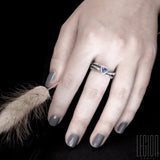 Woman's hand with gray nails wearing a black textured silver ring composed of 3 twigs crossing on the top and a blue tanzanite of triangular form set in close. 