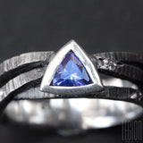 close-up of a black textured silver ring with 3 twigs crossing on top and a blue triangular tanzanite set in a closed setting with grey diamonds scattered on the ring body 